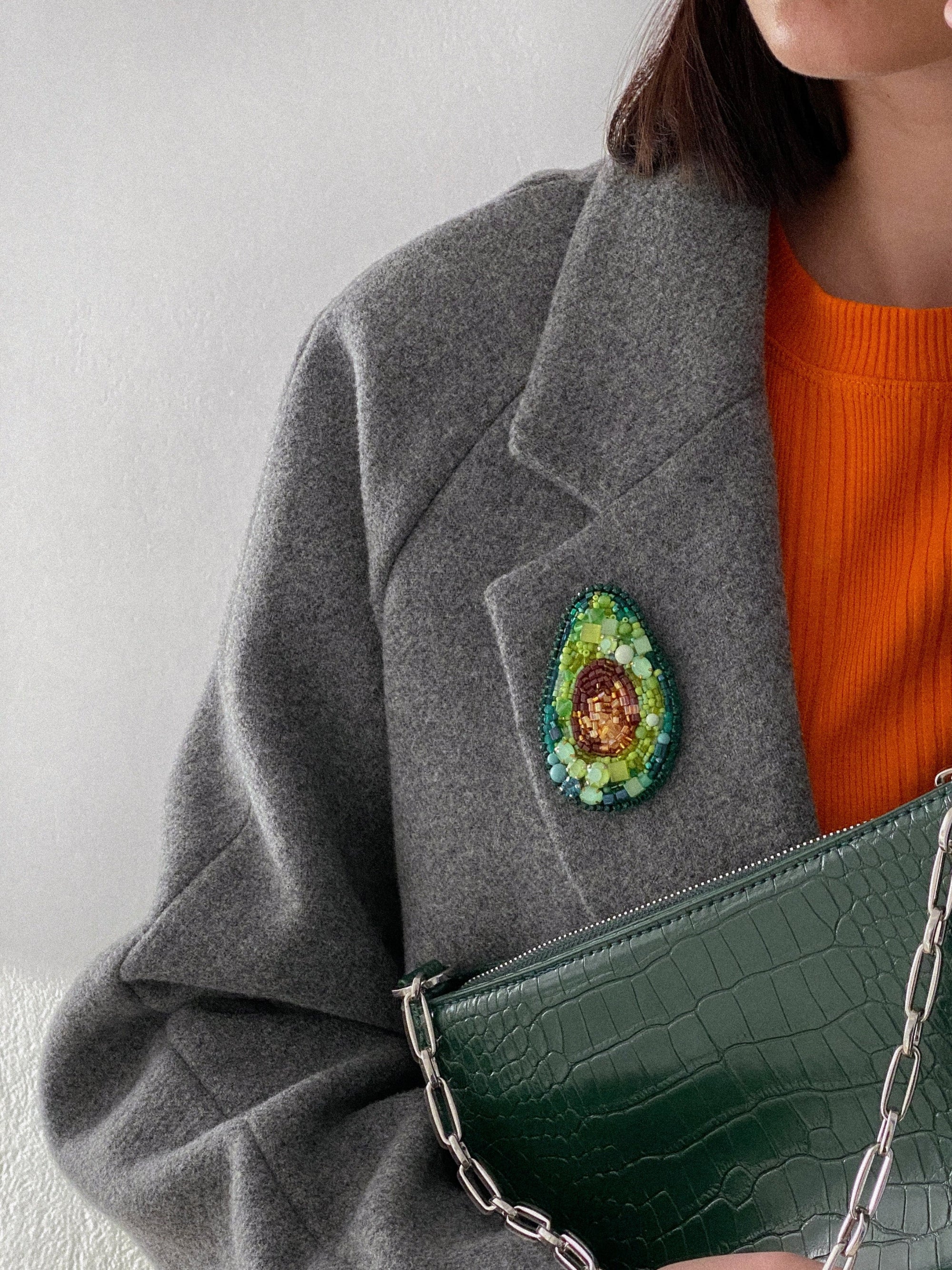 Person wearing a gray coat with an avocado brooch on the coat lapel, holding a green handbag.