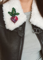 A close-up of a person wearing a black faux-leather jacket with a white sherpa collar, featuring a glittery green leaf and pink heart-shaped radish beetroot brooch pinned to the collar.