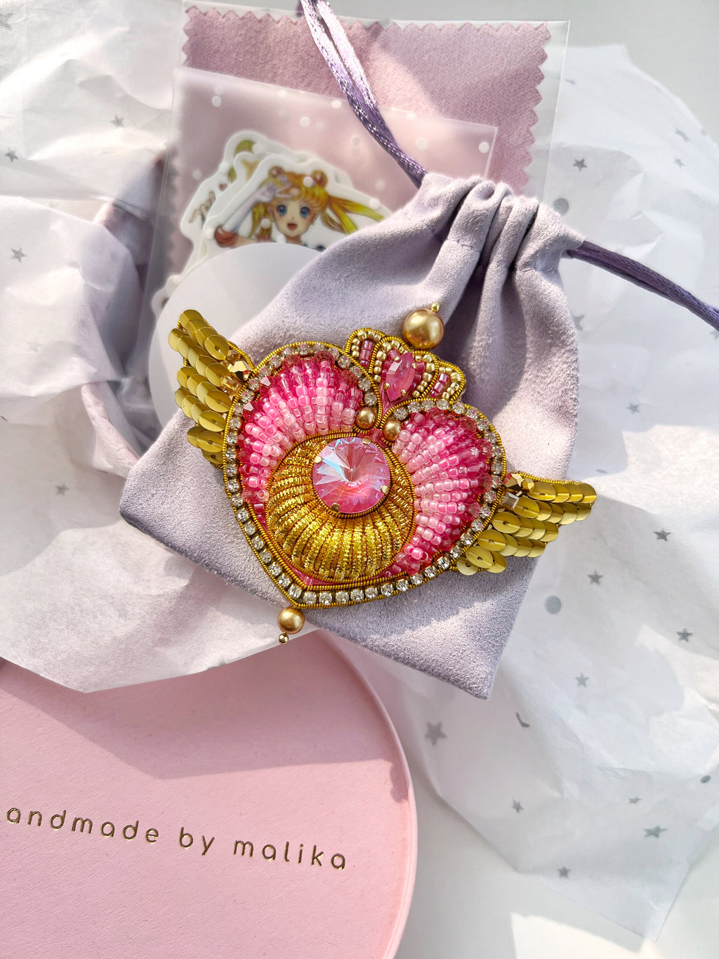 A decorative heart-shaped brooch or pin with pink and gold beadwork, angel wings, and a central faceted pink gem, displayed on a small lavender drawstring pouch with tissue paper and gift packaging around it.