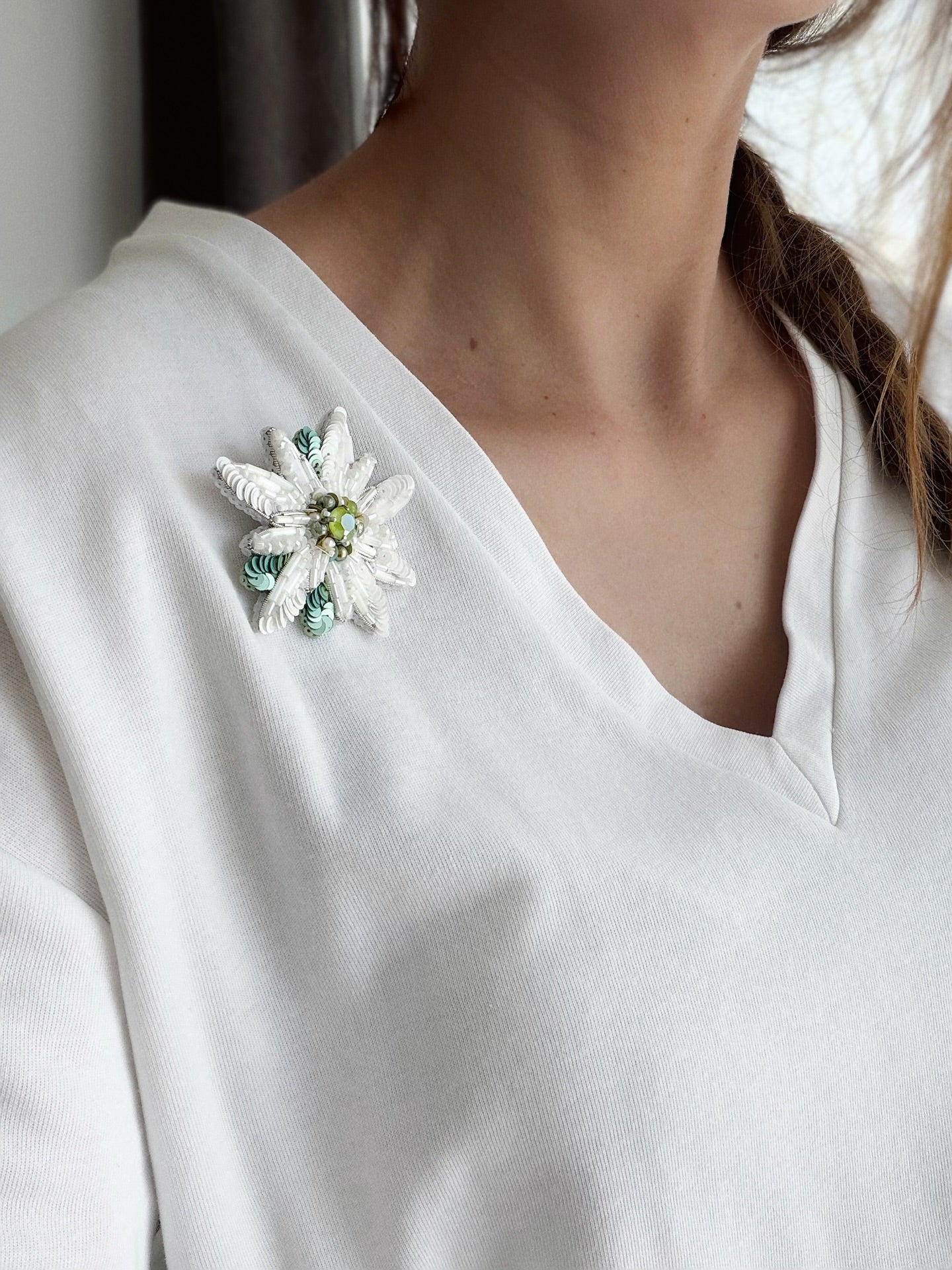 A close-up of a woman wearing a white V-neck top and a white flower edelweiss beaded brooch with green and yellow accents on her left shoulder.