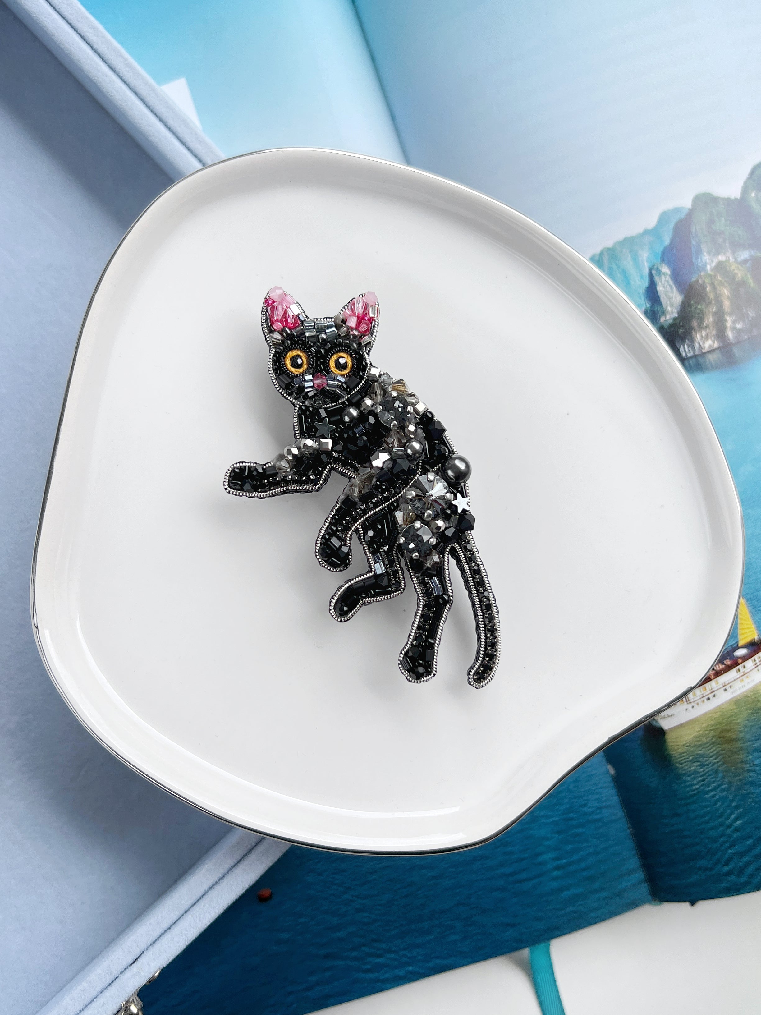 A black cat-shaped brooch with pink ears, yellow eyes, and silver rhinestone detailing, displayed on a white ceramic dish.