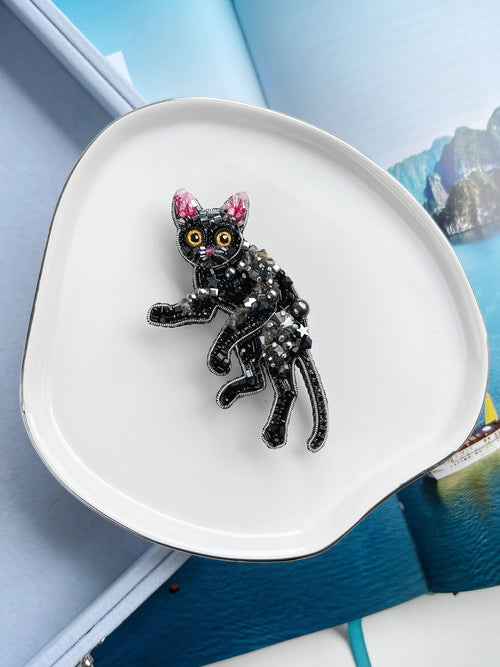 A black cat-shaped brooch with pink ears, yellow eyes, and silver rhinestone detailing, displayed on a white ceramic dish.