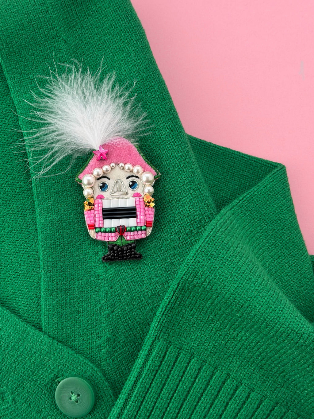 A close-up of a green knit cardigan with a pink nutcracker  brooch featuring a pink hat, white feather, pearl details