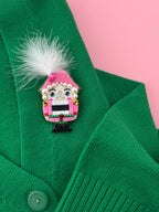 A close-up of a green knit cardigan with a pink nutcracker  brooch featuring a pink hat, white feather, pearl details