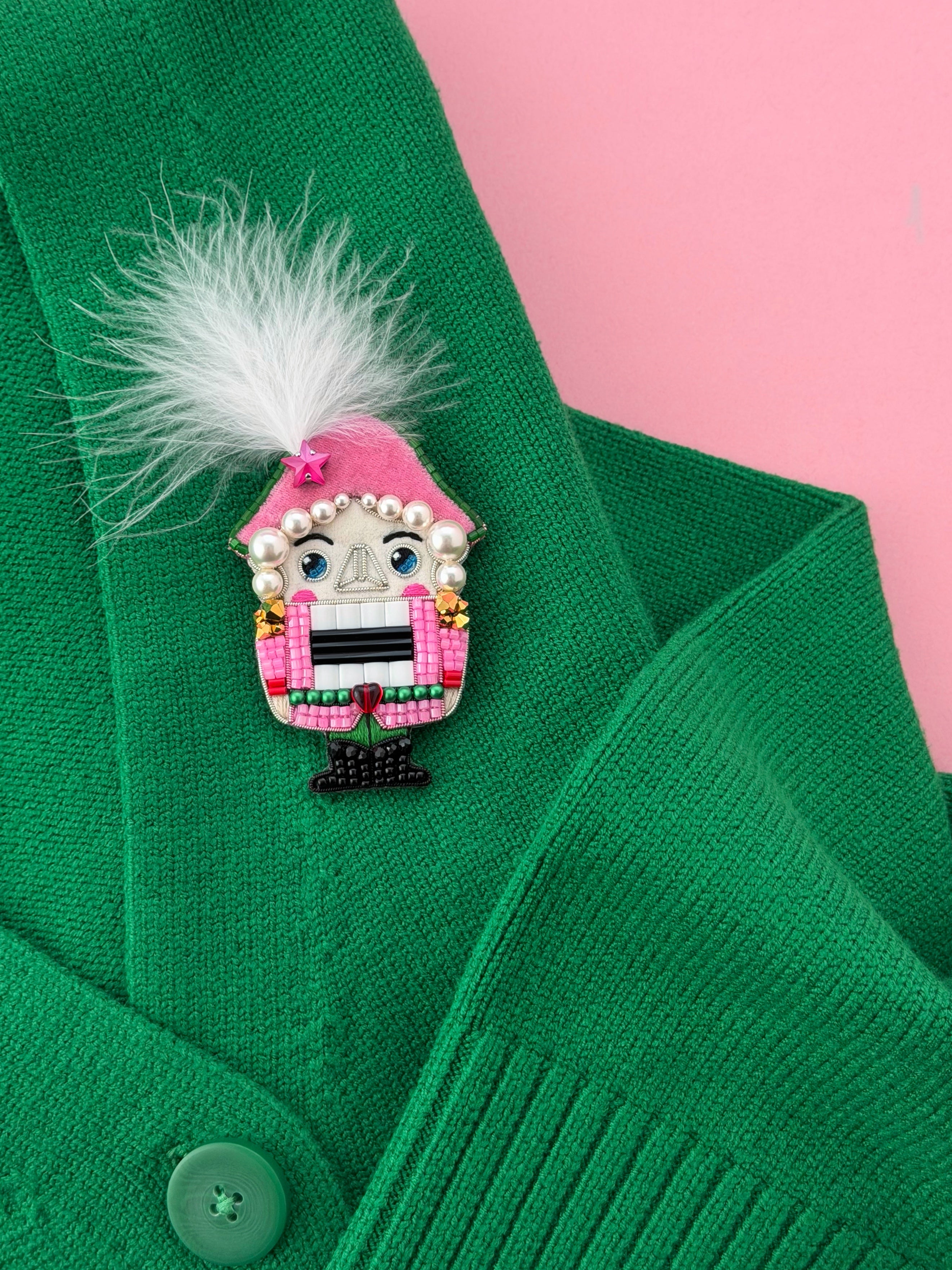 A close-up of a green knit cardigan with a pink nutcracker  brooch featuring a pink hat, white feather, pearl details