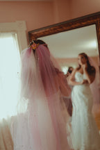 Woman in a white wedding dress with a pink veil fastened with Crisis Moon brooch standing in front of a mirror.