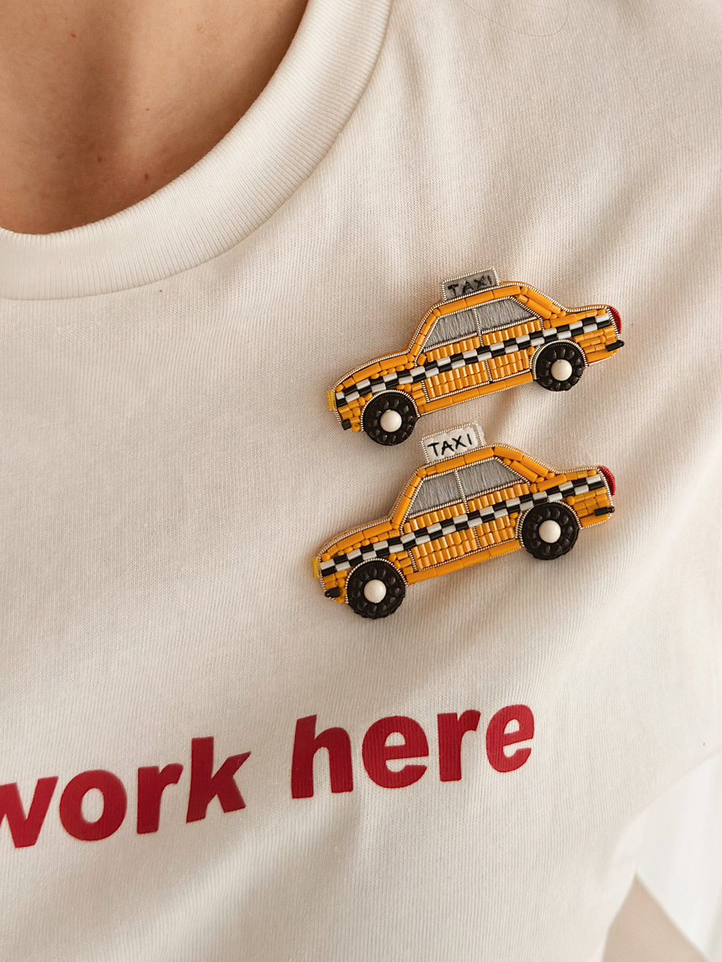 Two yellow and black checkered taxi-shaped pins pinned onto a white graphic t-shirt with red text.