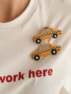 Two yellow and black checkered taxi-shaped pins pinned onto a white graphic t-shirt with red text.