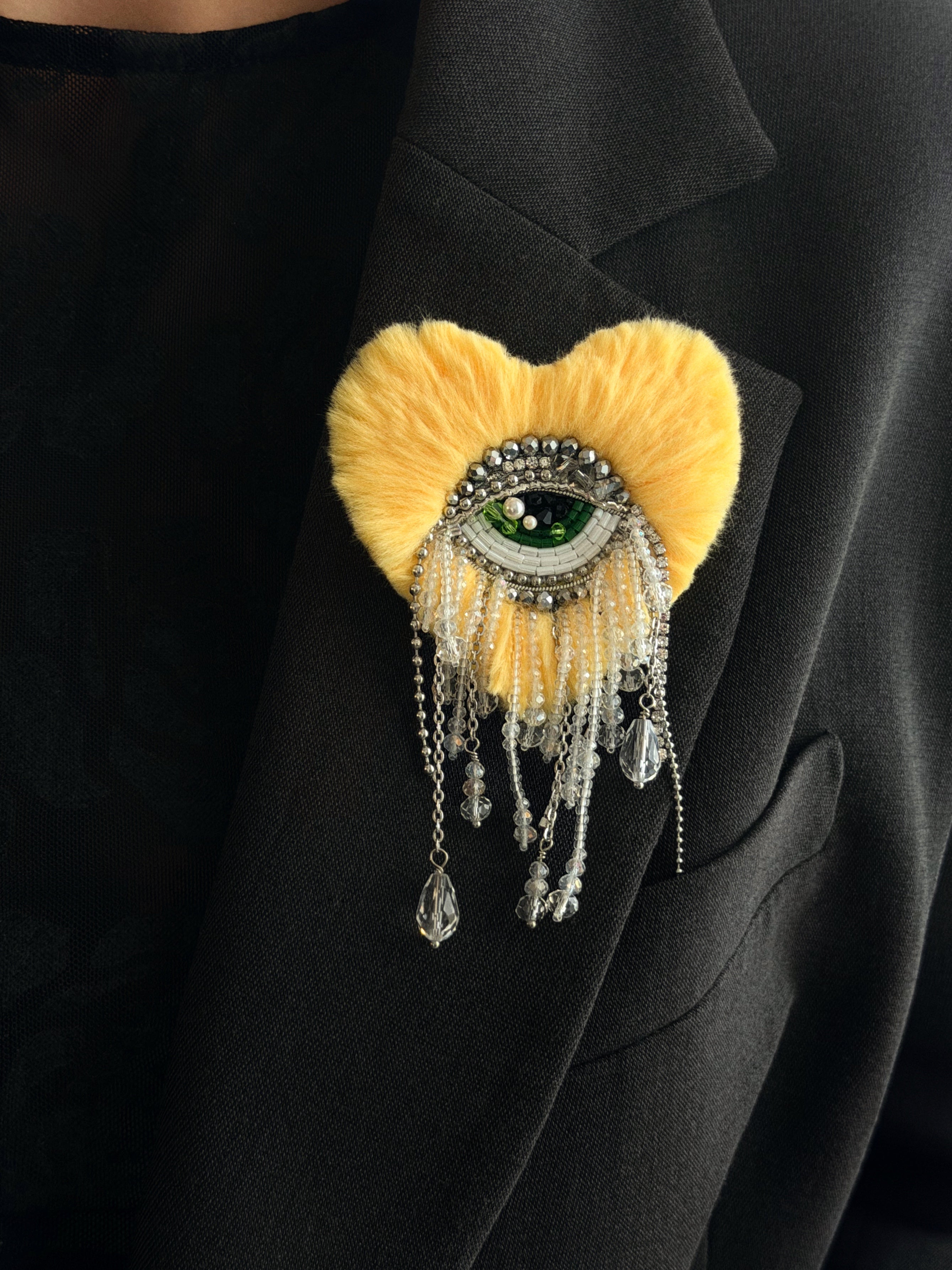 Yellow heart shaped brooch with eye design and dangling crystals on a black jacket.