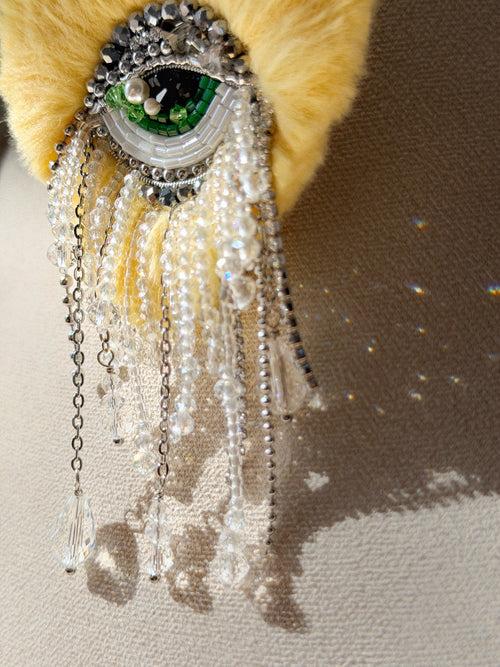Yellow heart with eye in the center brooch design, beads, and chain fringe drop sparkle on a neutral background