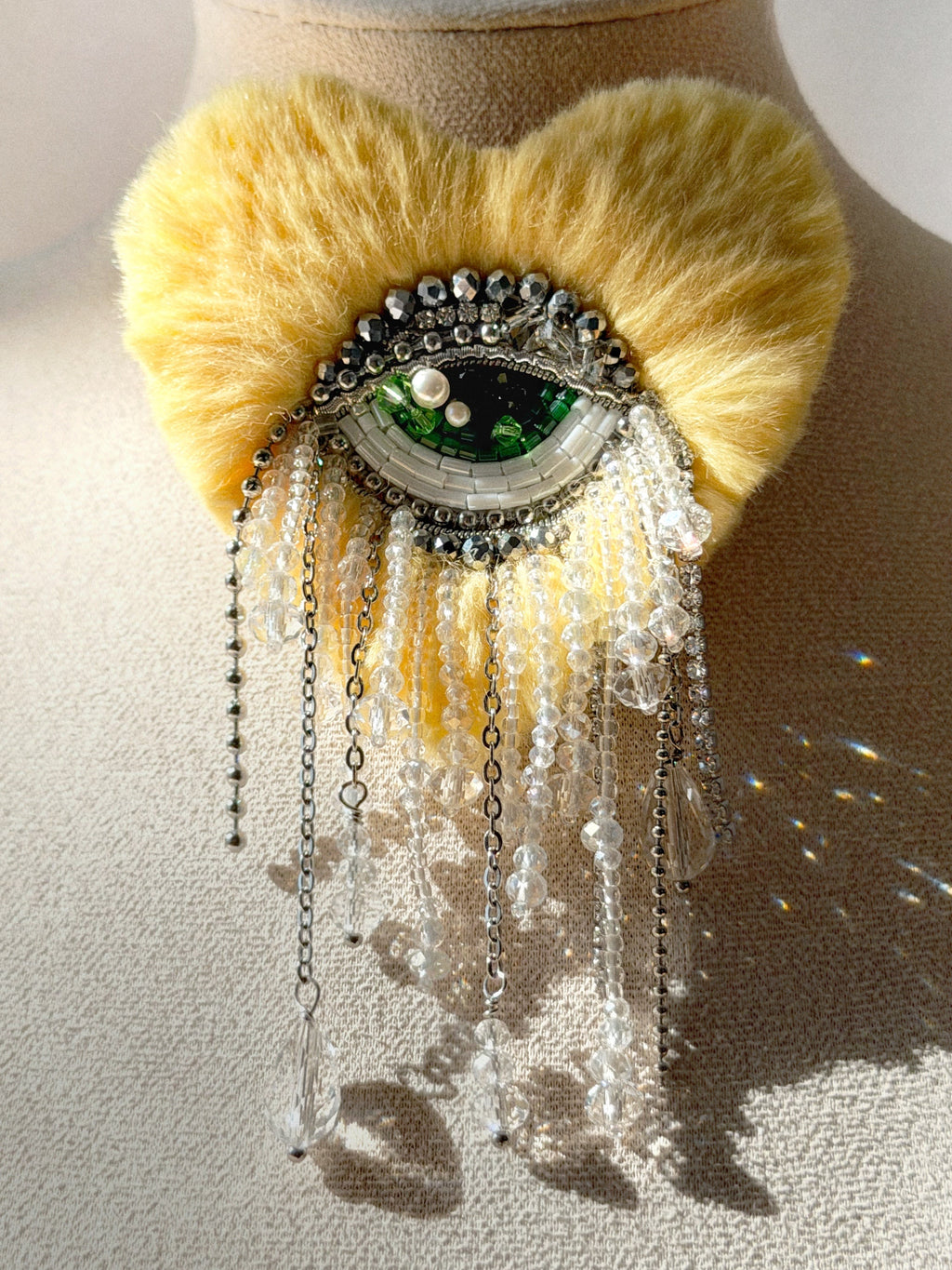 Heart shape brooch with light yellow  fur like texture and green eye design  in the center displayed on a mannequin bust.