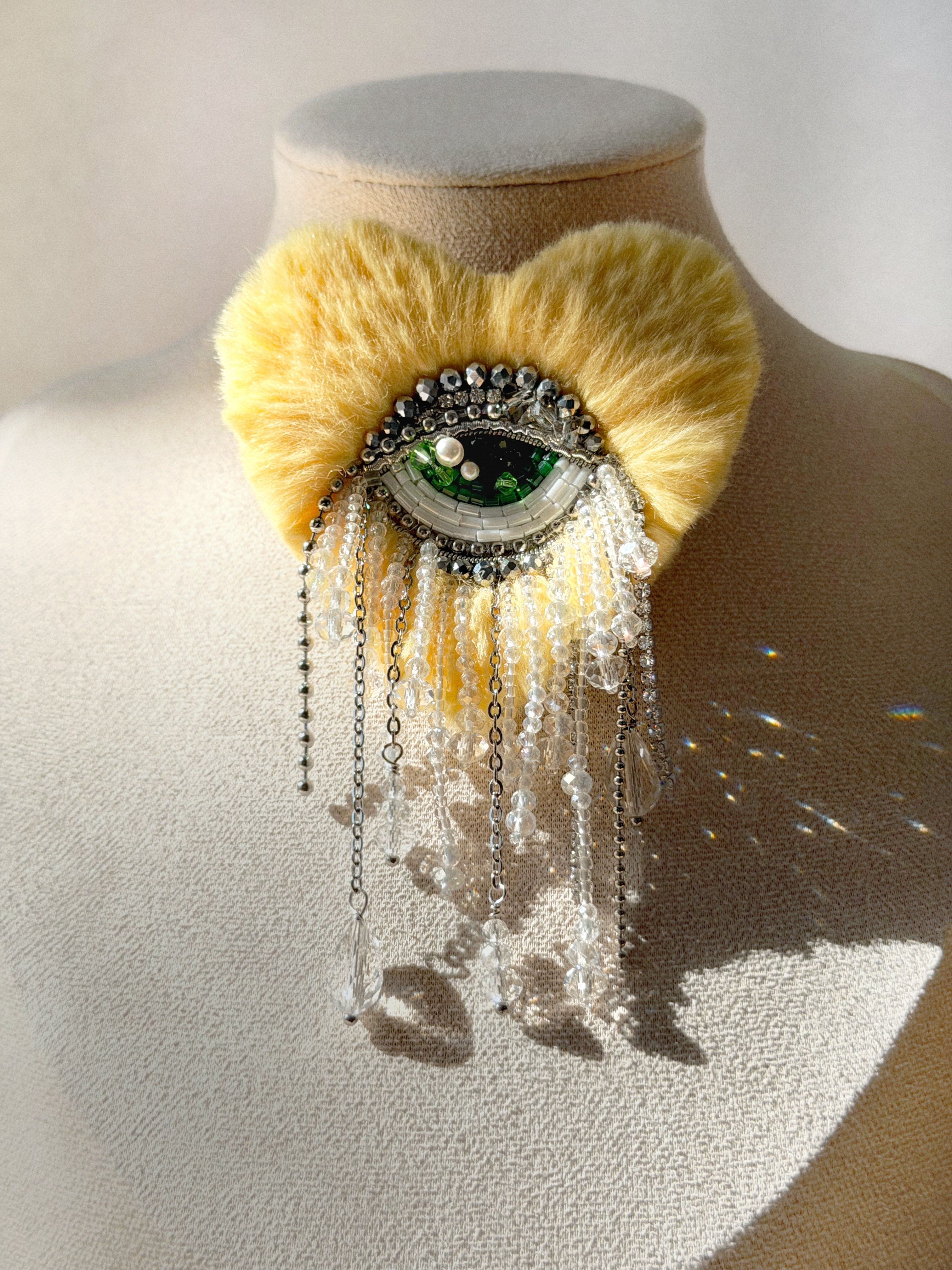Heart shape brooch with light yellow  fur like texture and green eye design  in the center displayed on a mannequin bust.